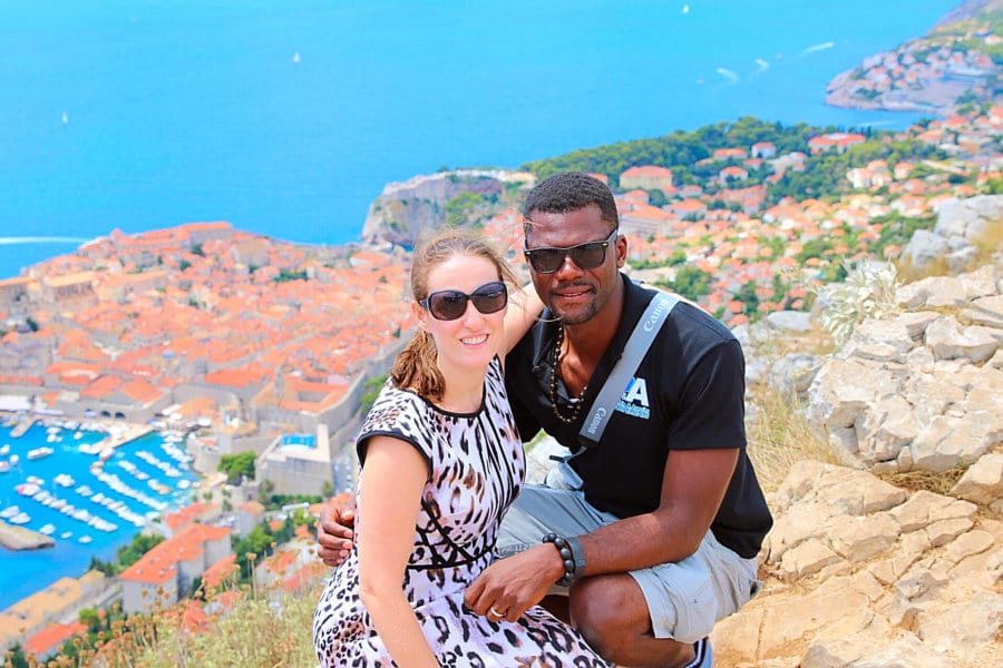 A couple smiles with the view of a town filled with red roofs near the ocean behind them.