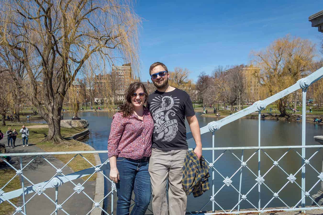A couple stands smiling on a bridge with the water behind them.