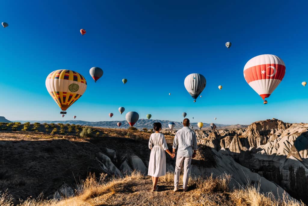 A couple holds hands while looking out at hot air balloons over mountains.