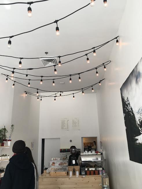 People stand in line to get coffee in a white room with string lights above.