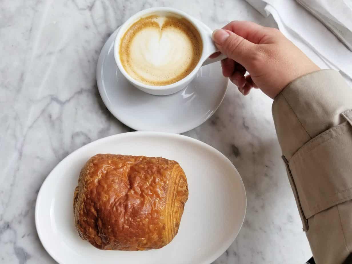 A person holds a white mug with coffee. A croissant sits on a plate.