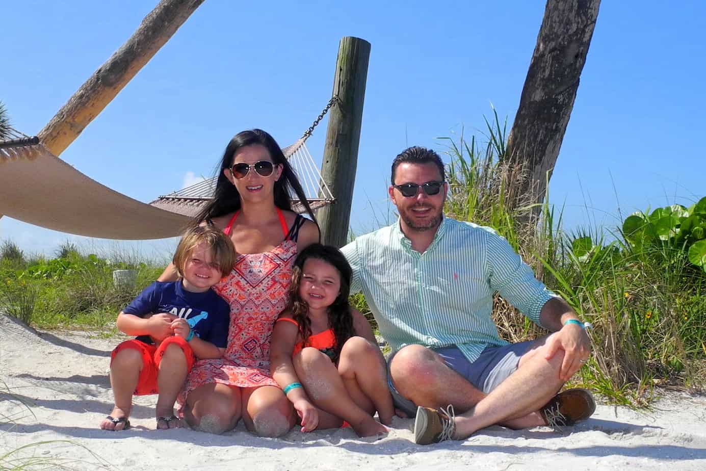 A family of four poses on the beach with a hammock behind them.