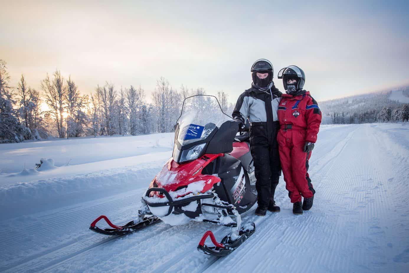 A couple in winter gear poses in the snow by a snowmobile