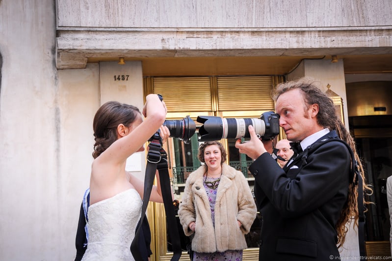 A couple in wedding outfits holds camera lenses to one another