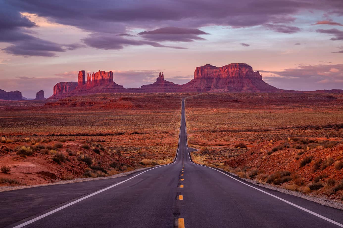 Empty road leading to giant red rock formations in the distance under a purple sky.