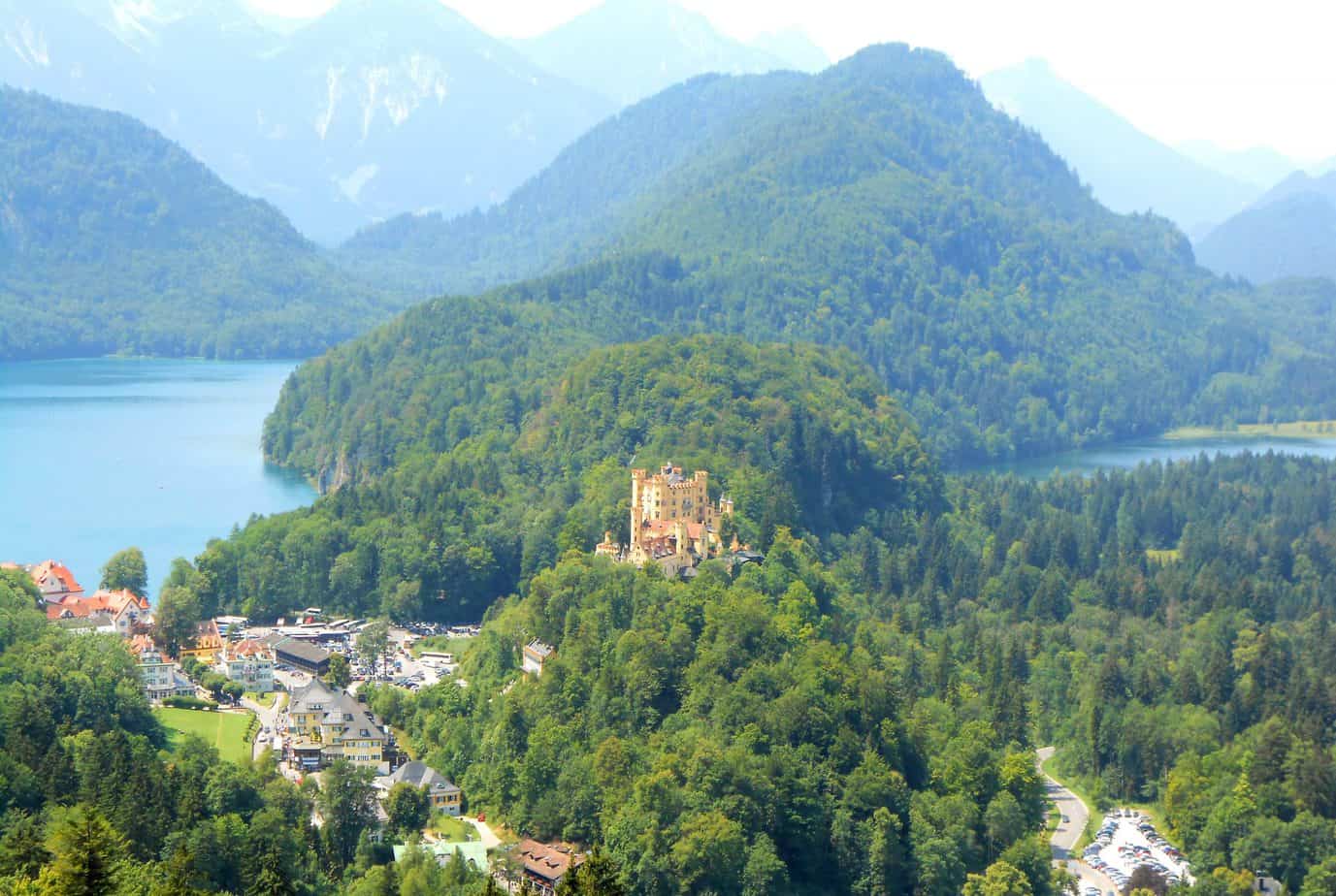 A small yellow castle is seen surrounded by mountains near a town and a lake.