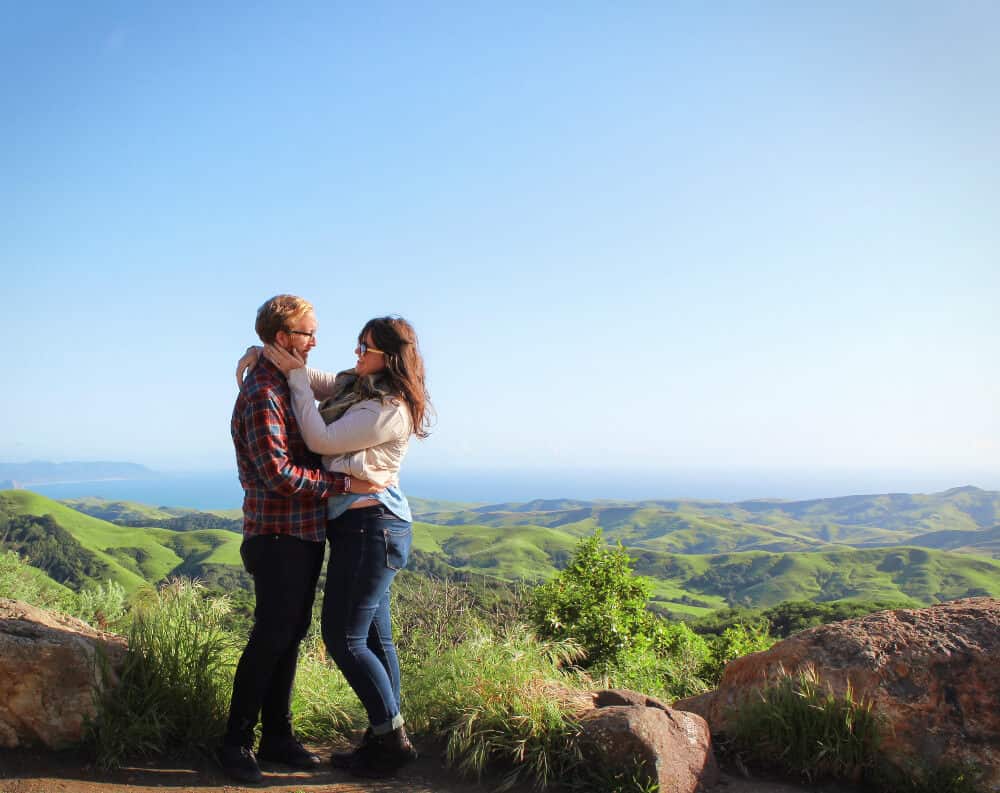 A couple holds one another under a blue sky with rolling hills in the background.