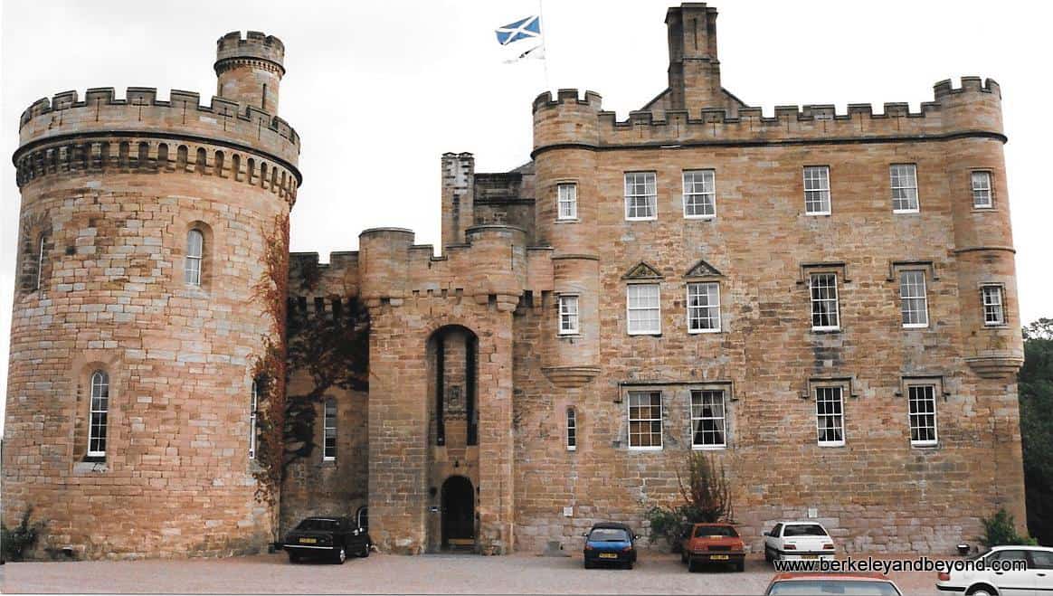A historic stone building with a Scottish flag on top. Cars are parked outside.