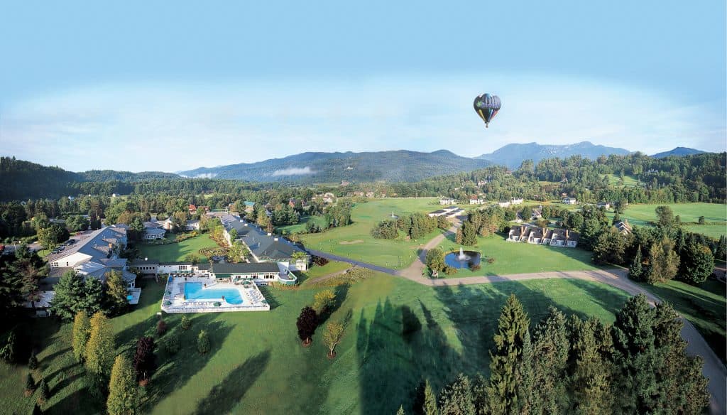 A hot air balloon floats above a town with green space. Mountains are in the background.