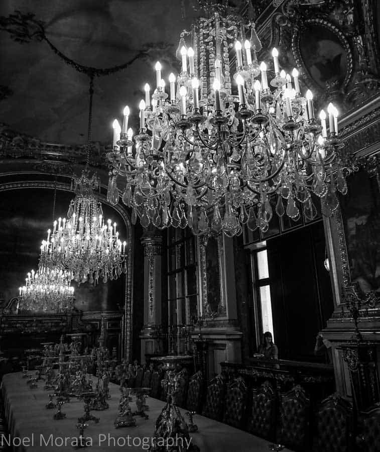 Chandeliers hang from the ceiling inside a palace in a black and white photo.