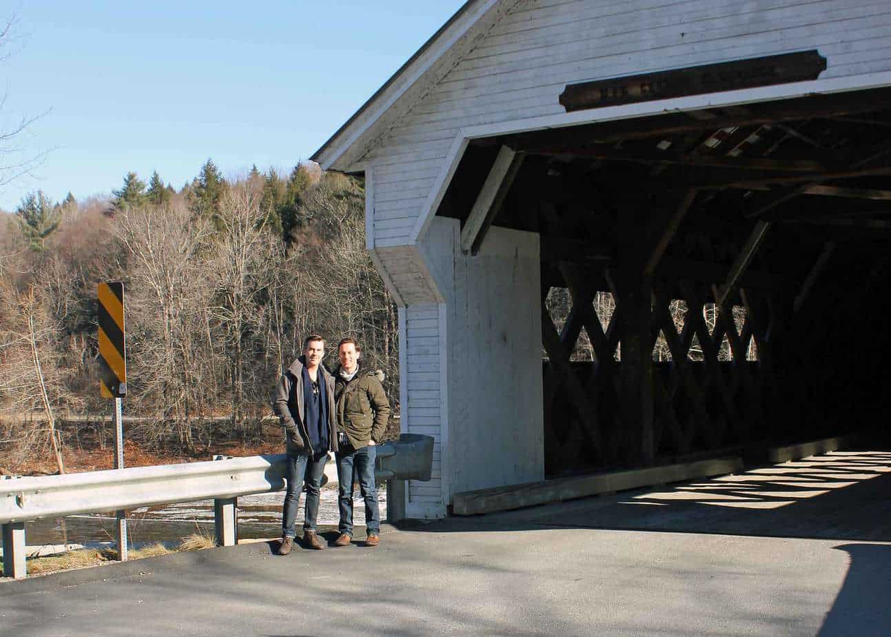 A couple poses in front of a white covered bridge.