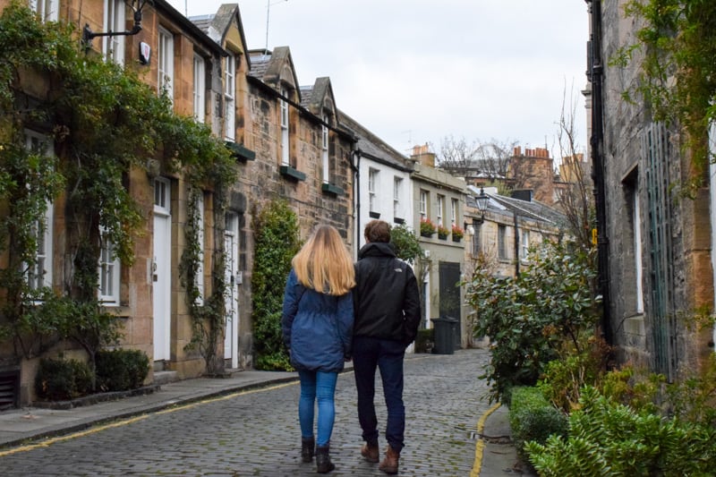 A couple walks hand in hand along a cobblestone street lined with buildings.