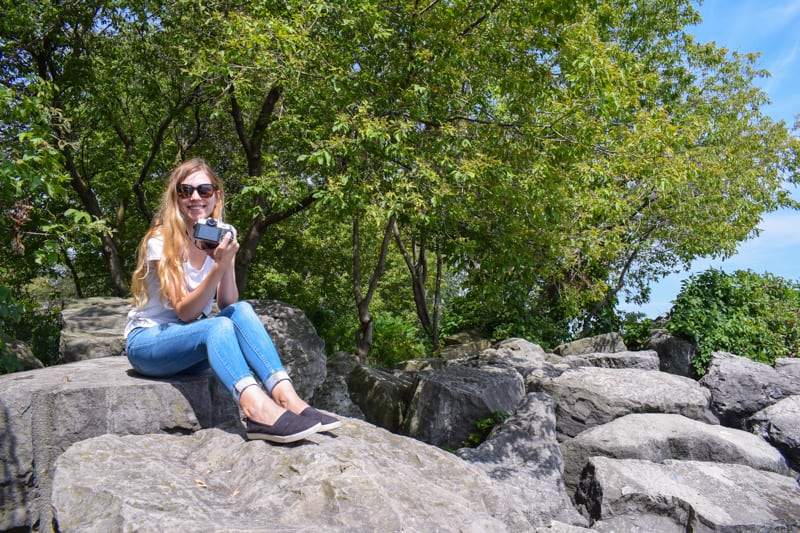A woman holds a camera while sitting on a rock.