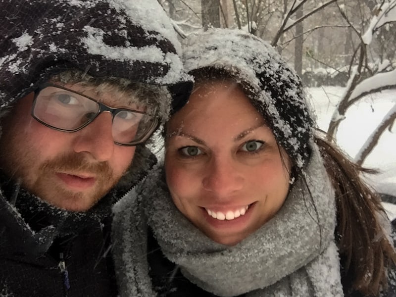 A couple smiles at the camera surrounded by snow.