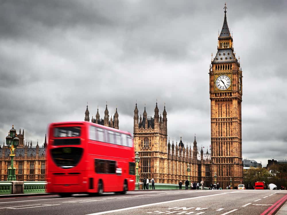 A red bus speeds past a London landmark on a grey sky day.