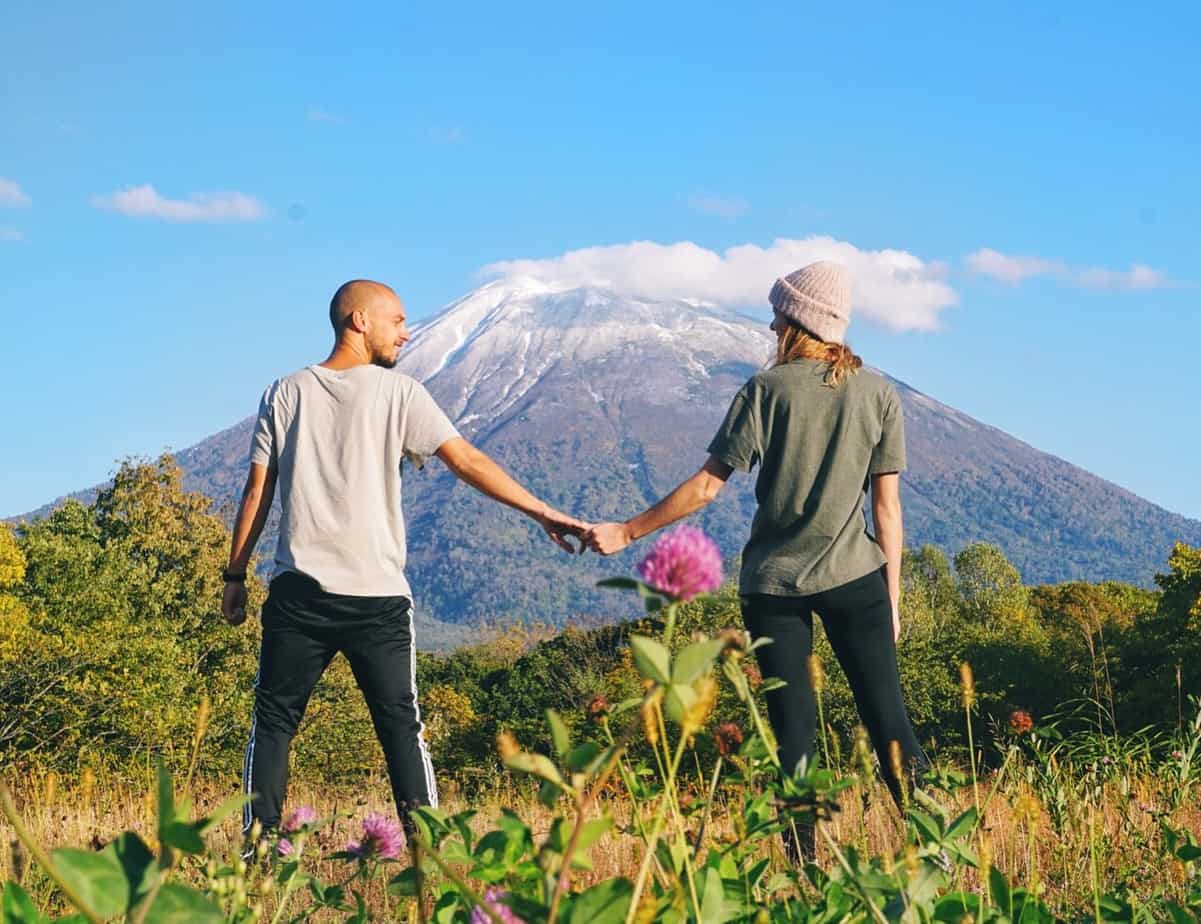 A couple holds hands in a field with a view of a snow-capped mountain behind.