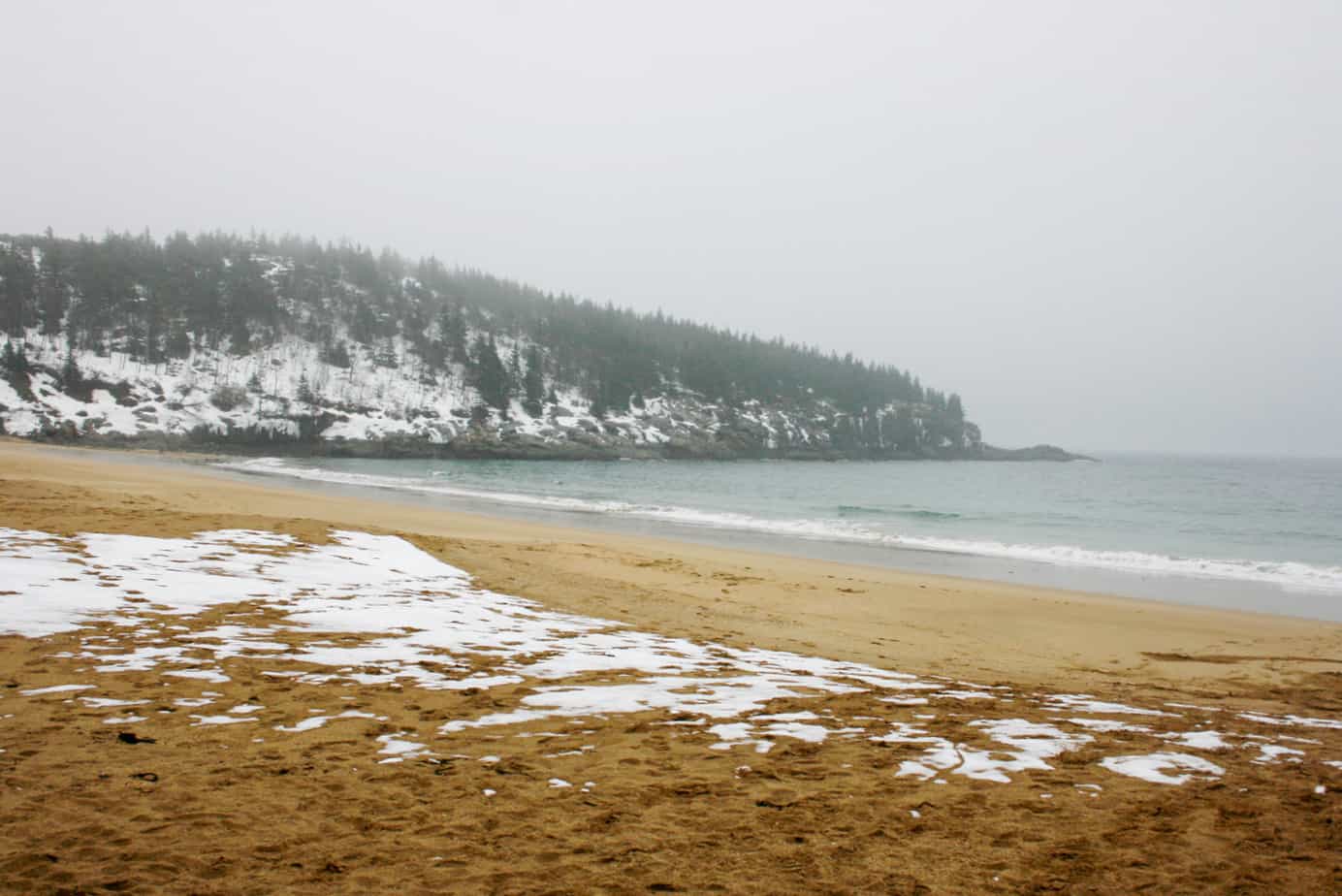 Waves crash against the coast on a foggy winter day. Some snow lines the mountain by the ocean, and the sand.