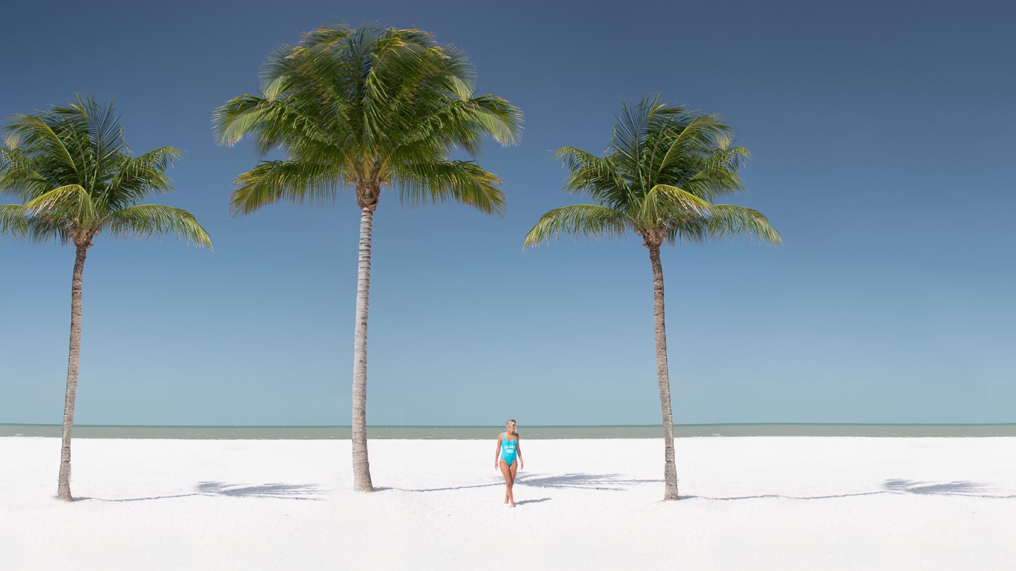 A woman walks below three palm trees on a white sandy beach