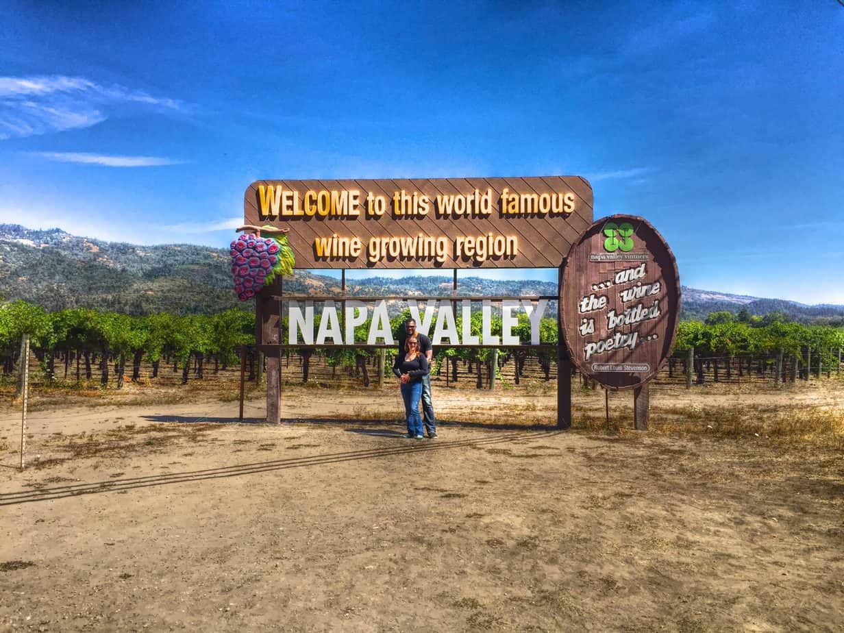 A couple poses in front of a sign that says \"Welcome to this world famous wine growing region. Napa Valley.\" with a vineyard behind them.