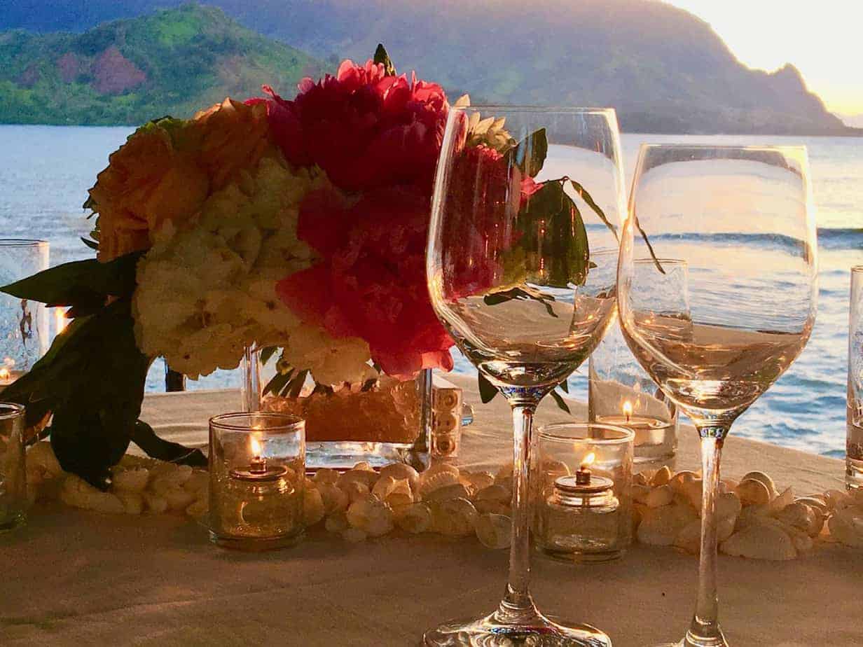 A flower arrangement sits on a table next to wine glasses with the ocean behind it.