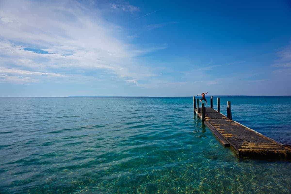 Long dock leading out into blue water with a person jumping off the edge. 