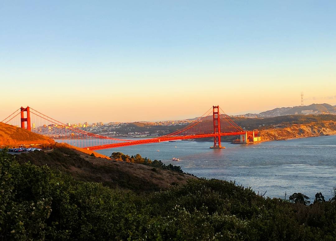 View of the Golden Gate Bridge from a viewpoint atop a hill in the distance at sunset. 