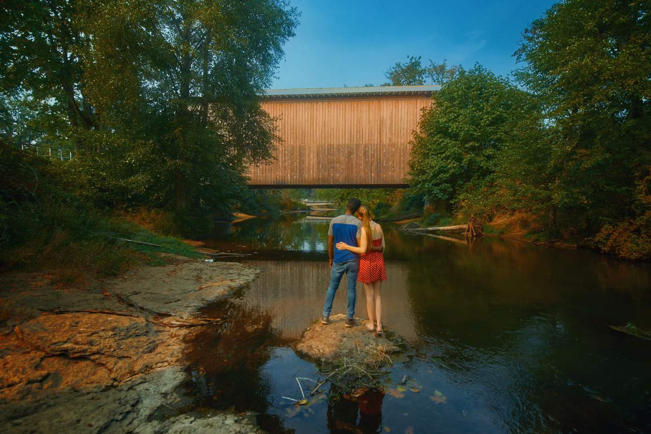 A couple looking at a covered bridge while standing on dirt in the middle of a river.