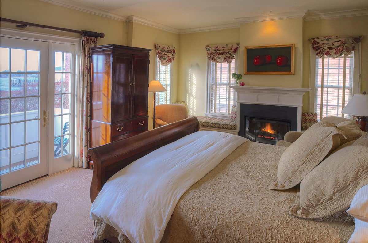 A bed is made with khaki-colored bedding. A fireplace is lit to the right.