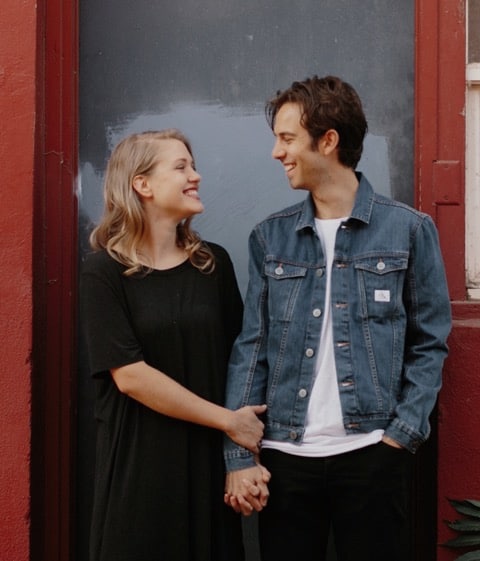 A man and woman smile outdoors. The woman is in a black dress and the man is in a denim jacket.