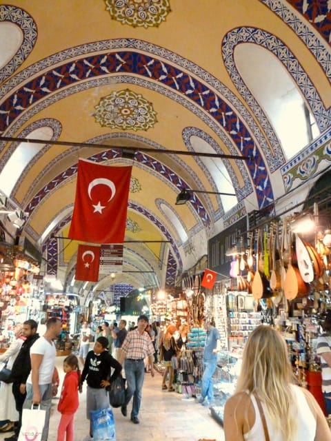 People walking around a grand bazaar filled with shops. Flags hang from the ceiling.