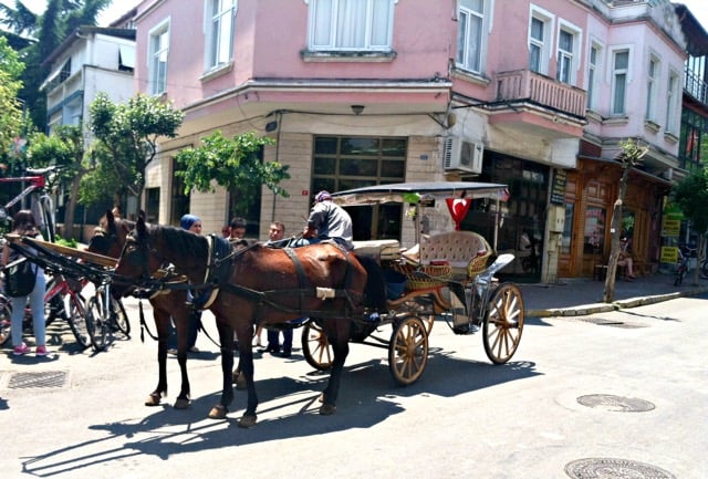 A man sits in a horse drawn carriage next to a pink building.