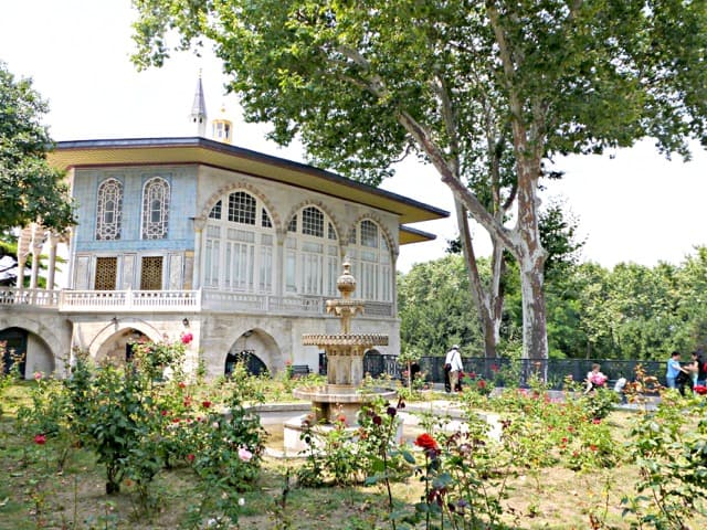 A romantic home with a garden in front featuring a water fountain. People are walking around the garden.