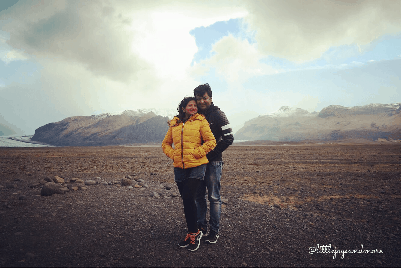 A man and woman standing next to one another in a dirt landscape with mountains behind them.