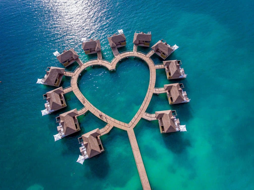 A heart shaped walkway leading toward bungalows floating in a bright blue sea. 