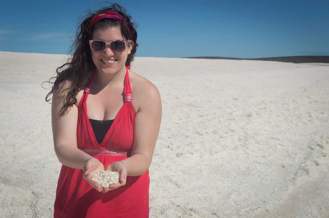 A woman stands smiling in a pink dress while holding sand on the beach