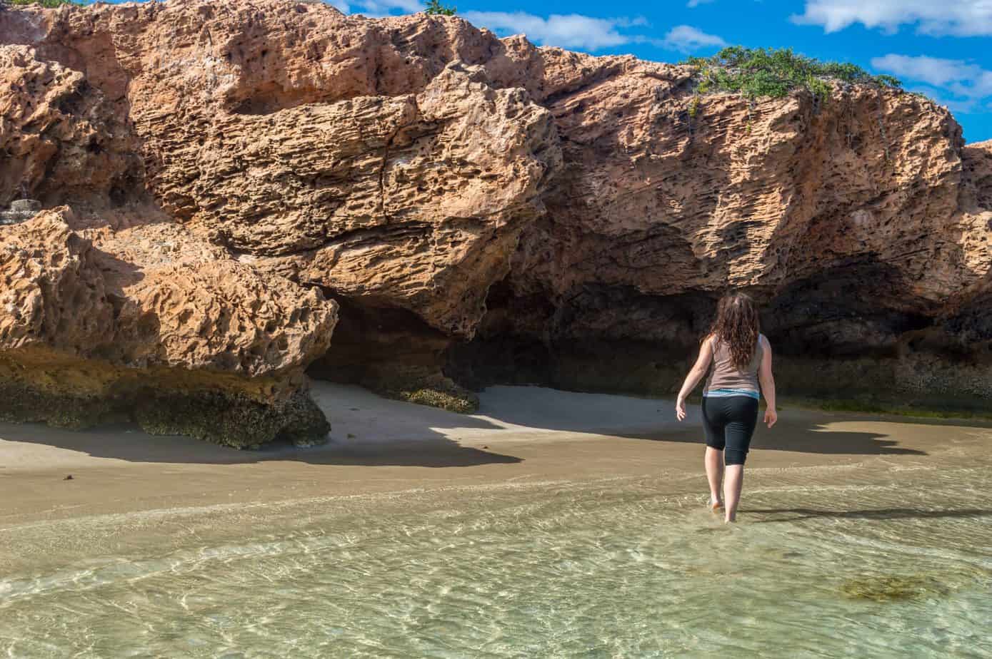 A woman walks in shallow ocean water next to a rocky cliff