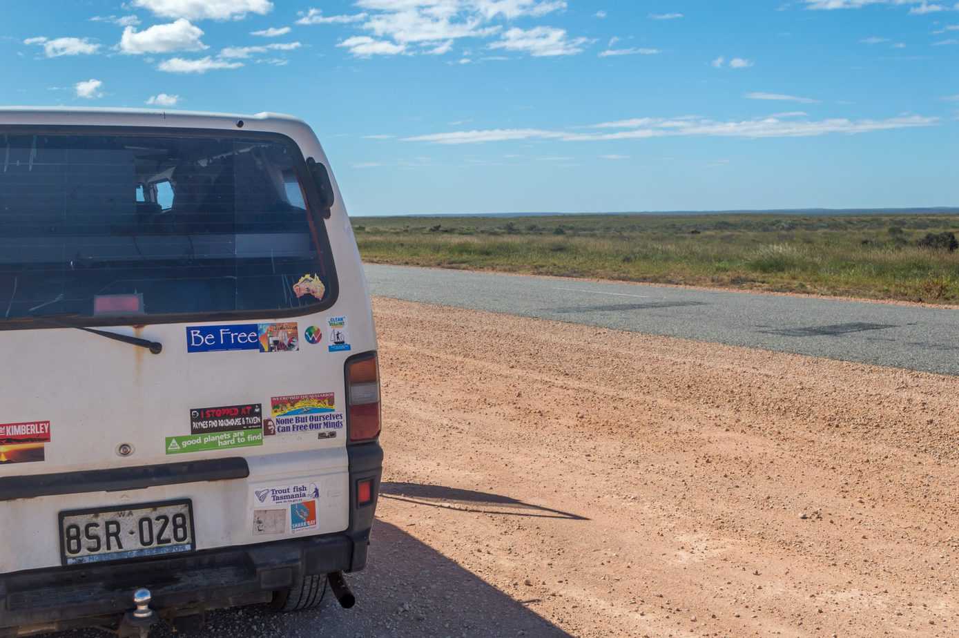 Back of a white van covered in stickers under a blue sky while driving along the road