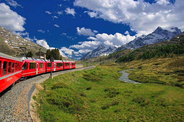 A red train travels through a landscape near a river. Snowy mountains are in the background.