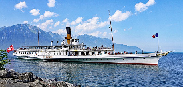 A large ship is packed with people on the water with a mountain behind.