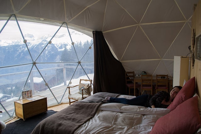 A person lays on a bed with views of snowy mountains.