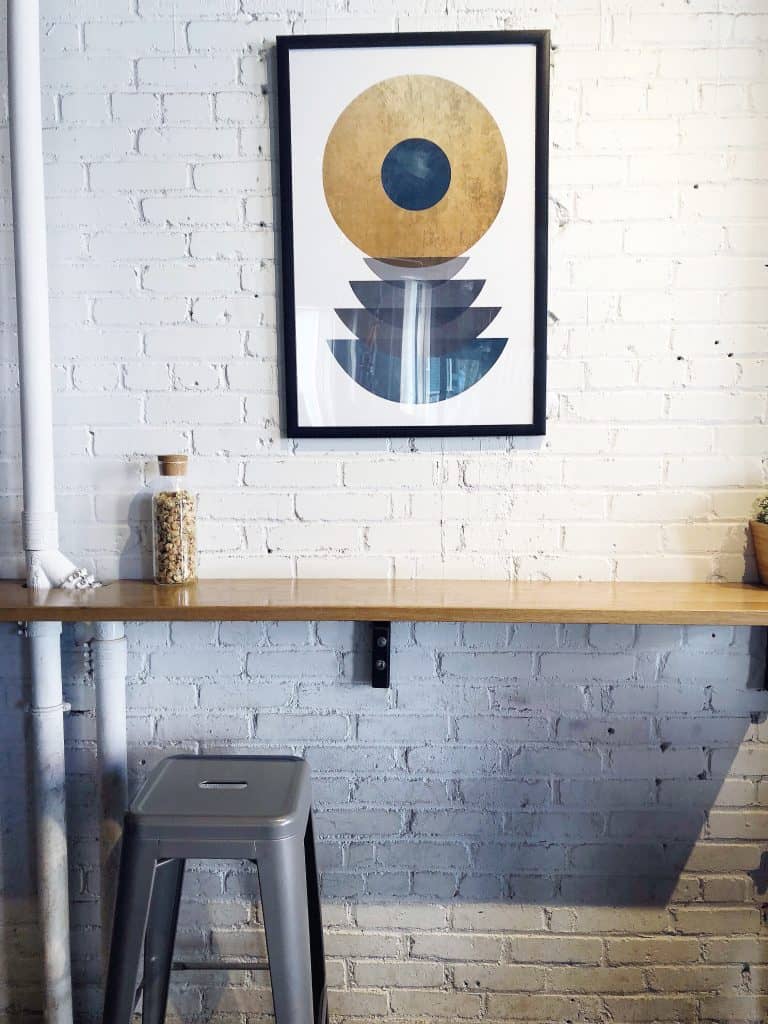 A grey stool sits next to a wooden table by a white brick wall. Above the table is a piece of art that\'s gold and blue with circular shapes.