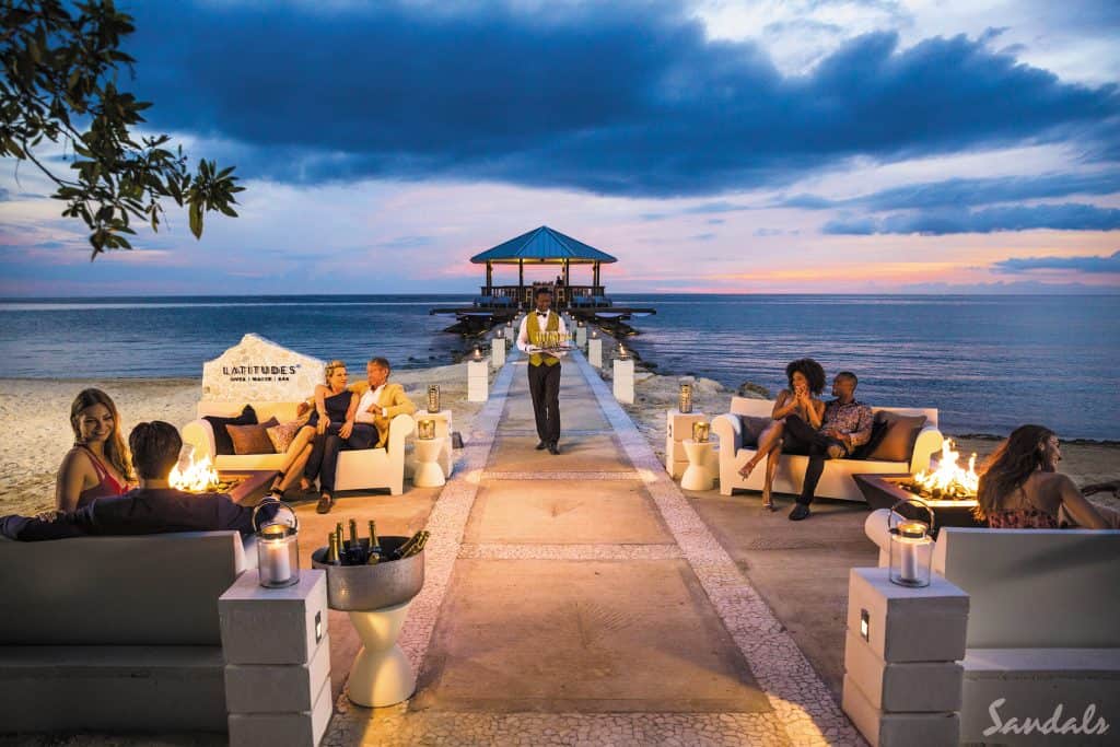 People sitting on couches on either side of a small pier leading out to blue water at sunset. 