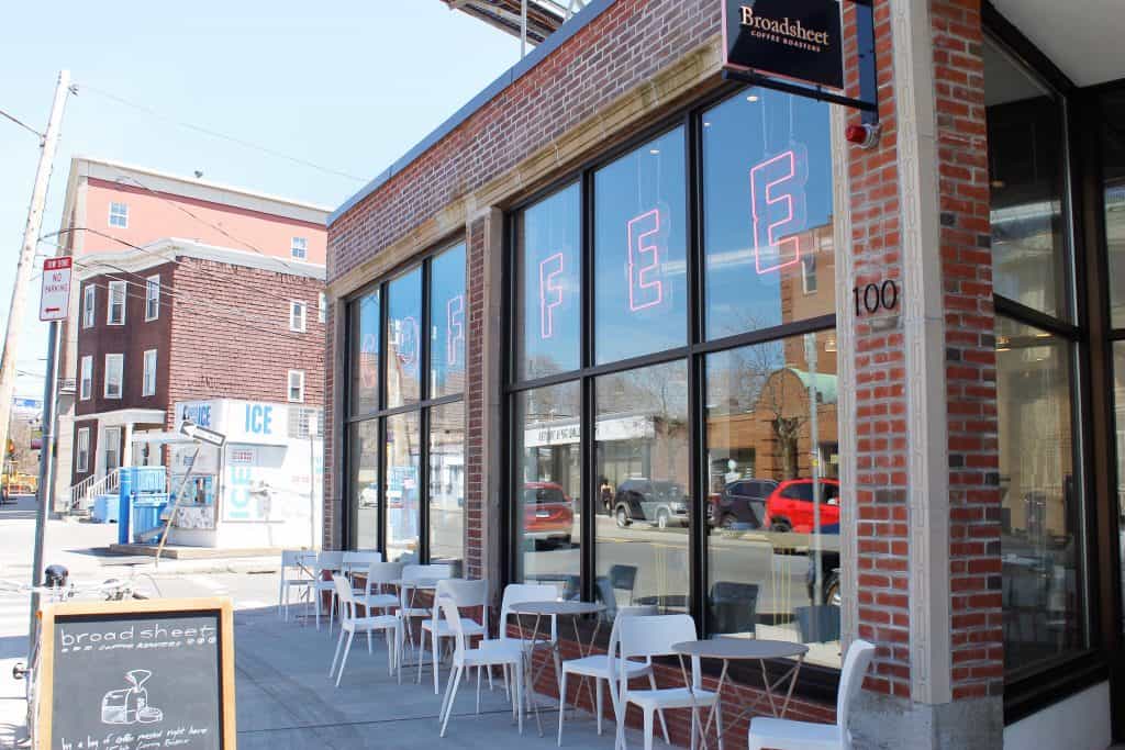 Exterior of a brick cafe with neon letters that spell out \"COFFEE.\" White tables and chairs line the sidewalk.