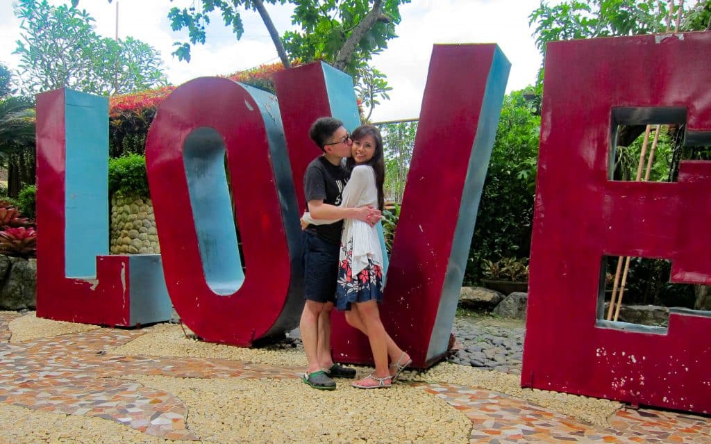 A couple kisses in front of a red sign that says LOVE.