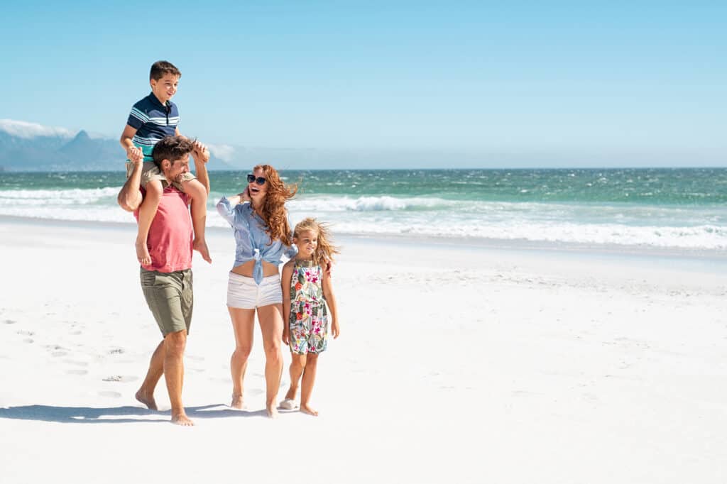 Smiling parents with children at sea. Cute son sitting on father shoulder with mother and sister walking while talking. Happy family with two children enjoying summer holiday at beach with copy space.