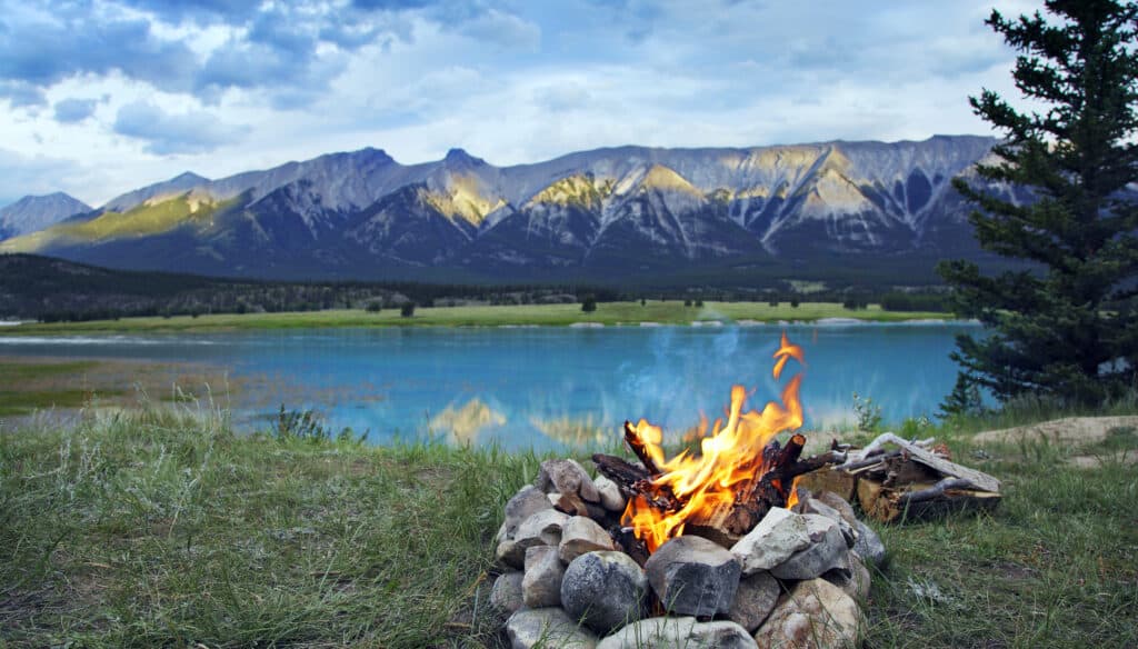 A fire surrounded by stones near a blue body of water and mountains in the background.