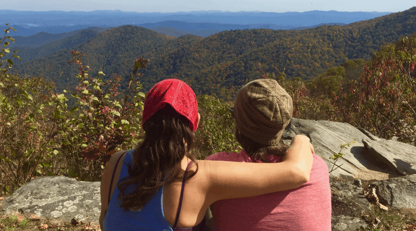 A couple has their arms around each other while looking out at mountains in green.