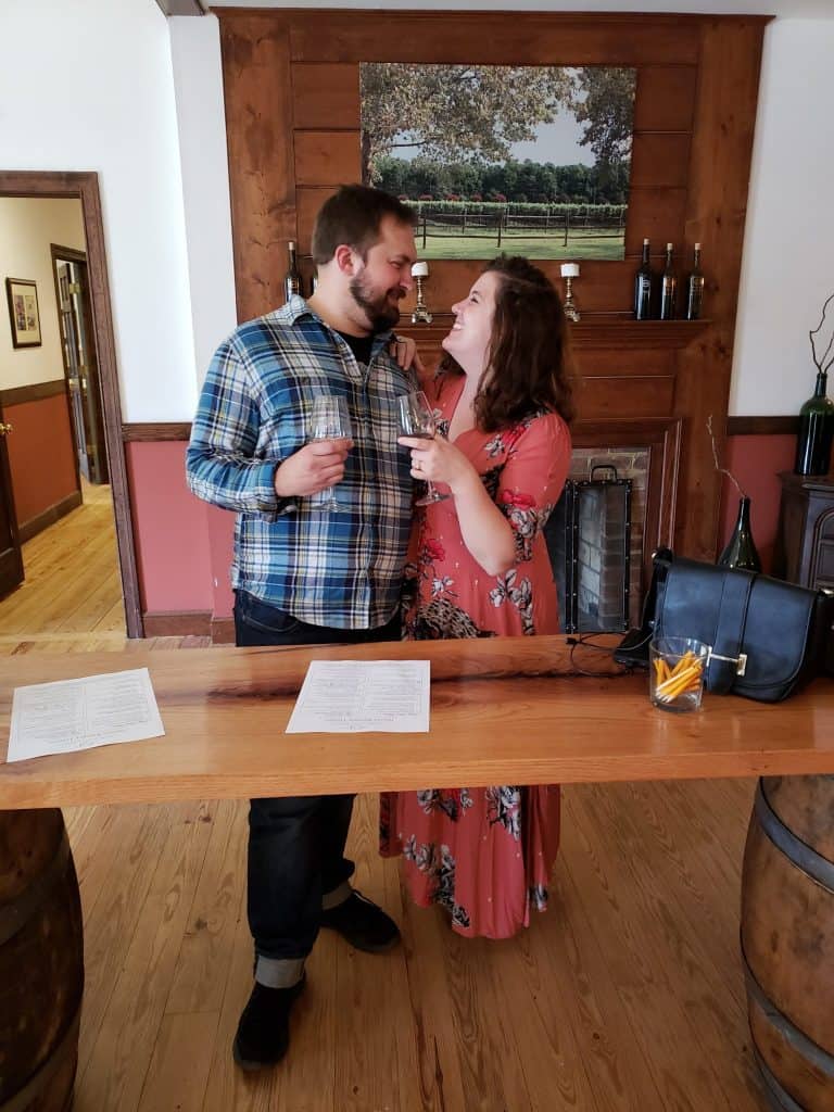 A couple smiles while holding wine glasses indoors.