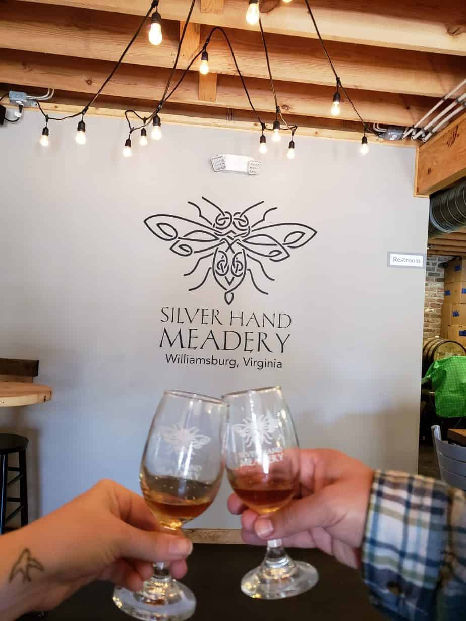 Two glasses of wine are being toasted in front of a white wall with writing that says "Silver Hand Meadery, Williamsburg, Virginia."