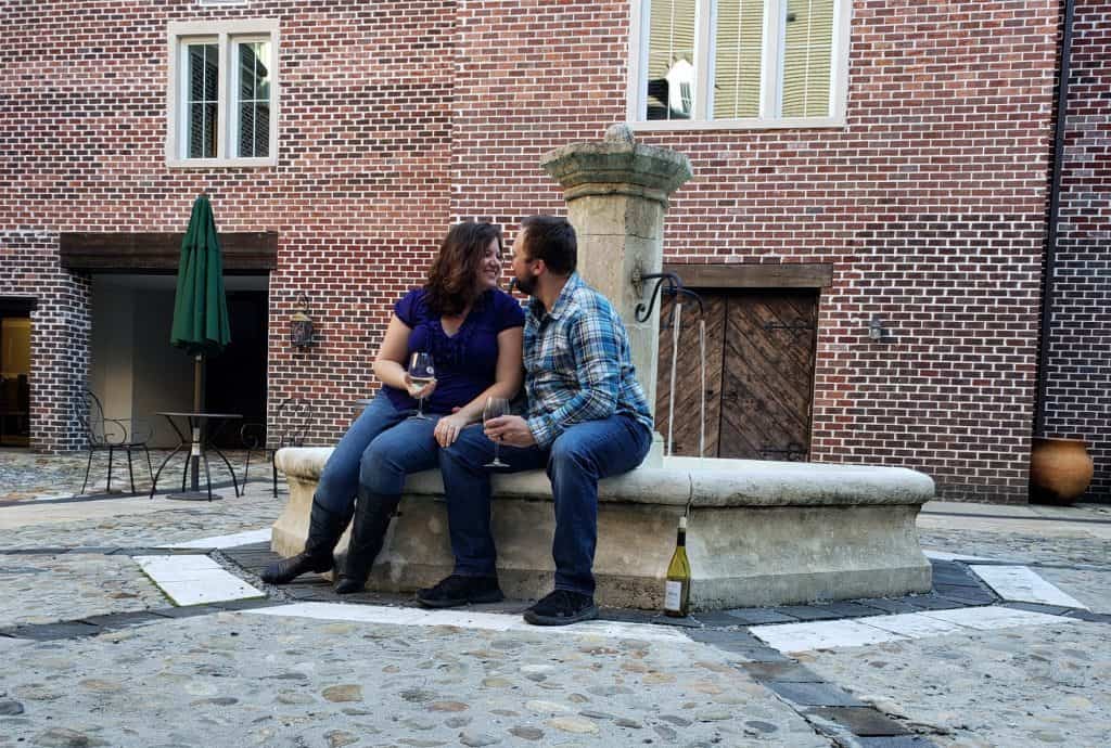 A romantic couple sits on the edge of a fountain with a bottle of wine at their feet. They are also each holding a glass of wine.